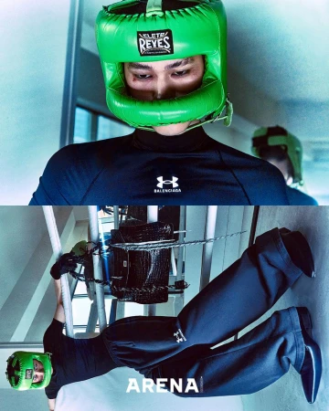 A person wearing a green boxing headgear, a black long-sleeved shirt, and black pants performs a pull-up on a metal bar in an indoor setting.