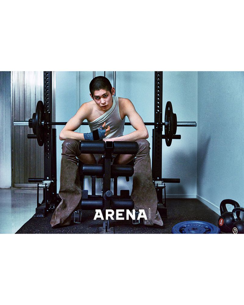 A muscular person poses on a weight bench with a barbell, dressed in a sleeveless top and distinctive brown pants, in a gym setting.