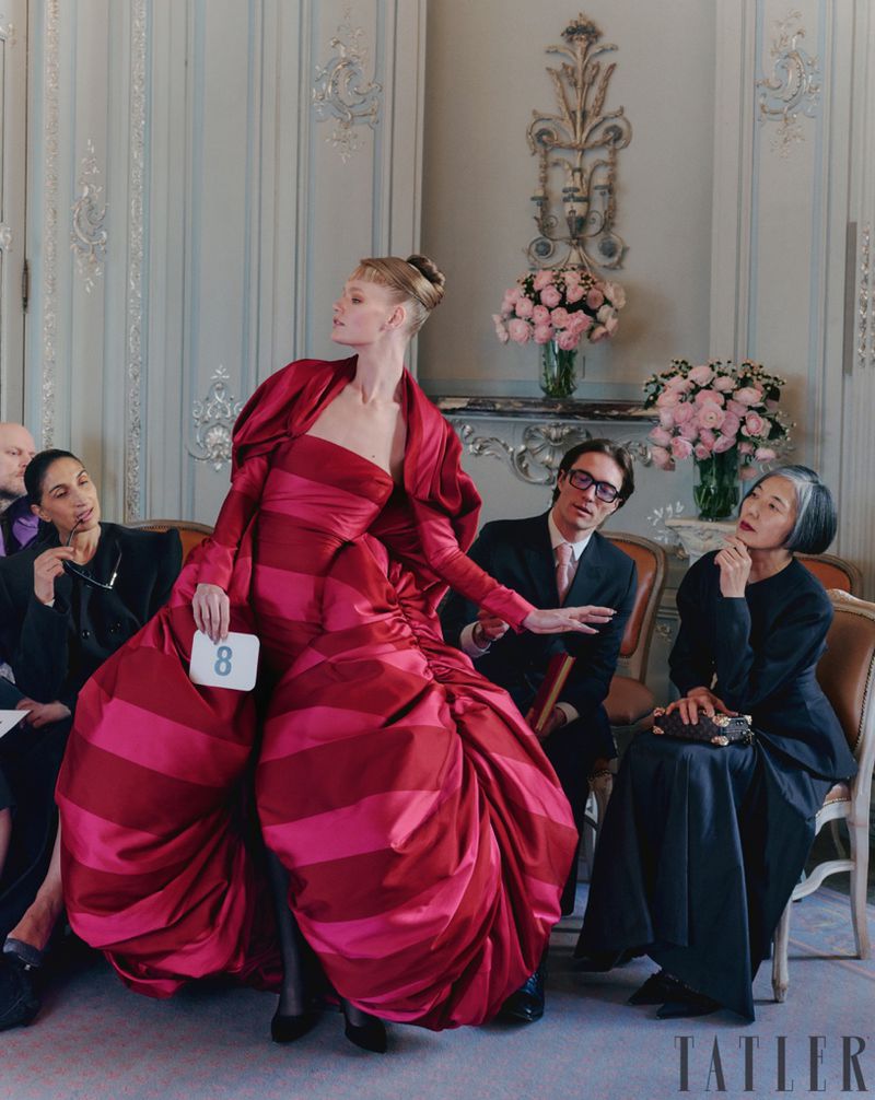 Fashion show featuring a model in a red gown with striped accents, seated observers, and floral decorations against an ornate wall.
