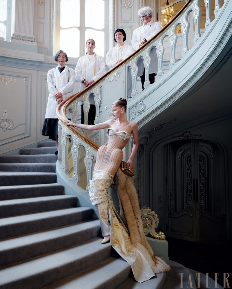 Fashion editorial shoot on a grand staircase, model in ornate dress, chefs in background, high-fashion elegance in a luxury interior.