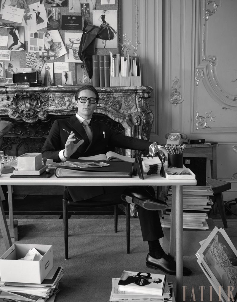 Black and white shot of a man in a suit seated at a desk in an ornate room, with books, papers, and a fireplace visible. 