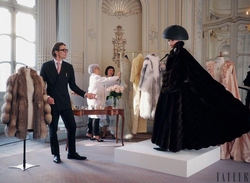 Fashion shoot in ornate room features a model in dark fur coat and hat, with two women adjusting a garment and a man in suit.