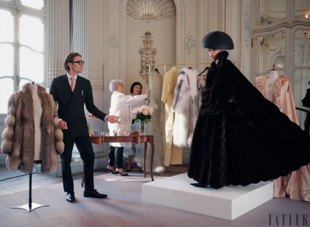 Fashion shoot in ornate room features a model in dark fur coat and hat, with two women adjusting a garment and a man in suit.