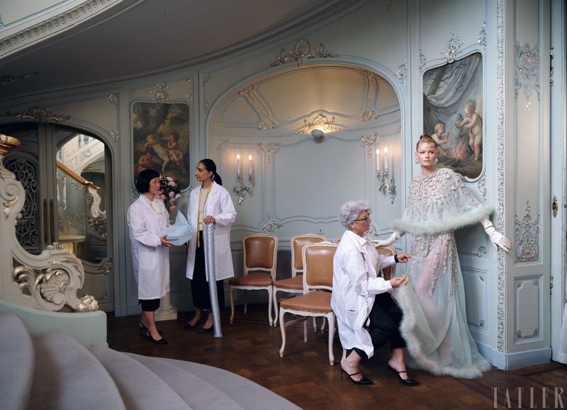 Fashion editorial shot indoors with a model in a couture gown being attended to by three women in white coats, art nouveau environment.