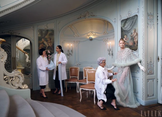 Fashion editorial shot indoors with a model in a couture gown being attended to by three women in white coats, art nouveau environment.