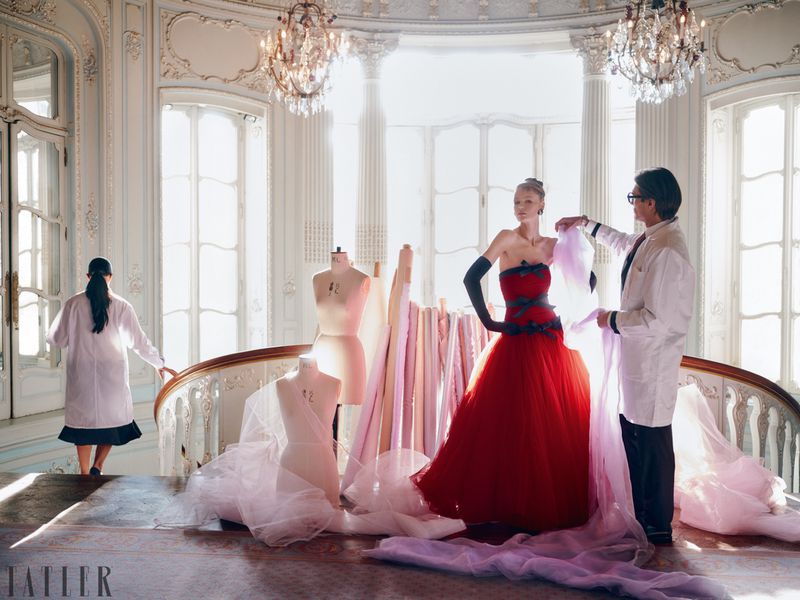 Fashion editorial featuring a model in a red gown, with a designer adjusting the fabric in a grand, light-filled hall with architectural details.