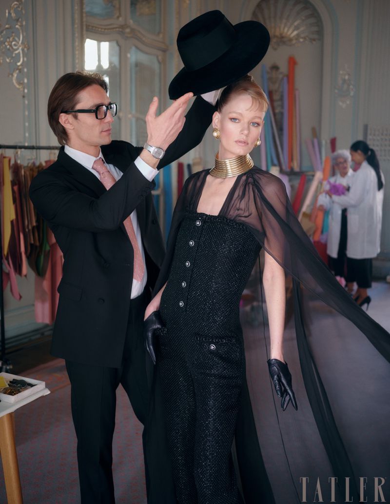 Model in a black dress and a designer adjusting a hat on her head in a fashion studio, featuring a neutral indoor setting.