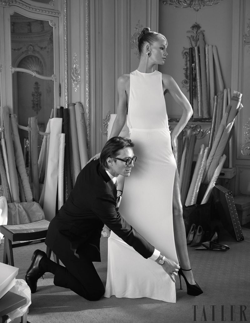 A fashion designer adjusts the hem of a white dress worn by a tall model in a photo studio with rolls of fabric and ornate decor.