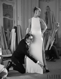 Black and white editorial fashion image of a man kneeling to adjust a woman in a long white gown standing in a cluttered atelier.