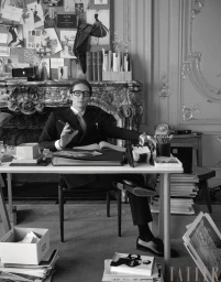 Monochrome image of a man in glasses seated at a desk in an ornate room with a corkboard and fireplace behind him