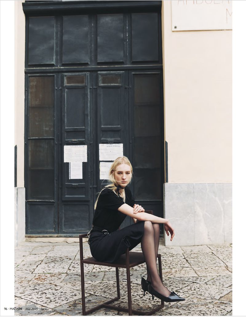 A blonde woman in dark clothing poses while seated on a metal chair in front of a large, black, paneled door outdoors.