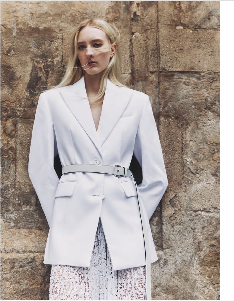 Fashionable woman poses in white blazer with belt against stone wall, creating a chic outdoor editorial look with natural light.