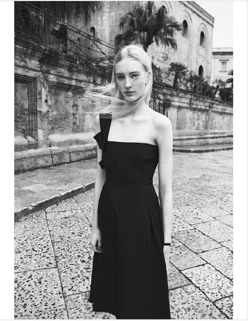 Monochrome outdoor portrait of a woman in a black dress on a cobblestone street with a vintage building and palm trees in the background.