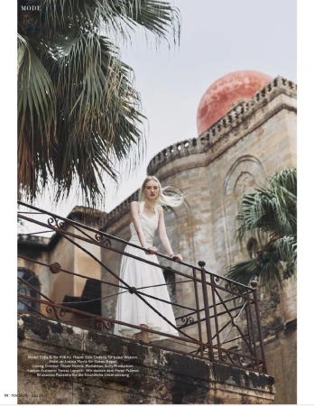 Woman in white dress stands on balcony in front of stone building with red dome and palm trees in a fashion editorial style shot.