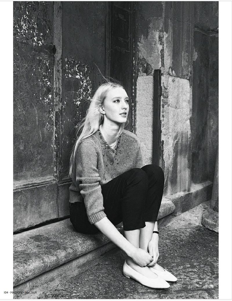 Black and white portrait of a blonde adult female sitting on a stone ledge with a textured wall in the background, looking upward.