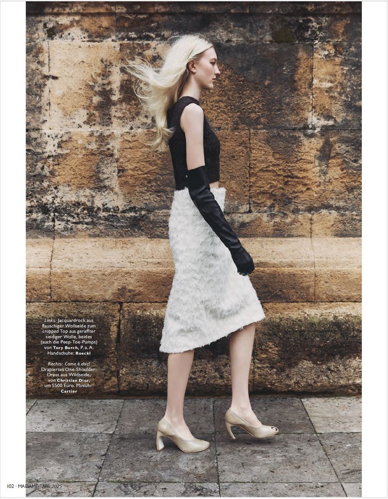 A blond woman models a black crop top, white skirt, and long gloves on a stone walkway, showcasing high-fashion style with a neutral backdrop.