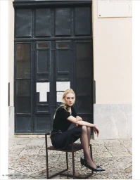 Model wearing a black dress and heels seated on a metal chair in front of a dark double door exterior for Madame Germany
