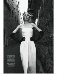 Model in white strapless dress and long black leather gloves posing in a narrow stone alleyway from a low angle
