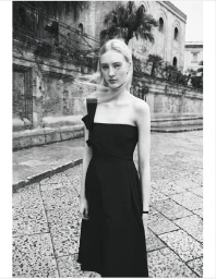 A blonde model wearing a black one-shoulder midi dress stands on a stone-paved street in front of an old stone building.