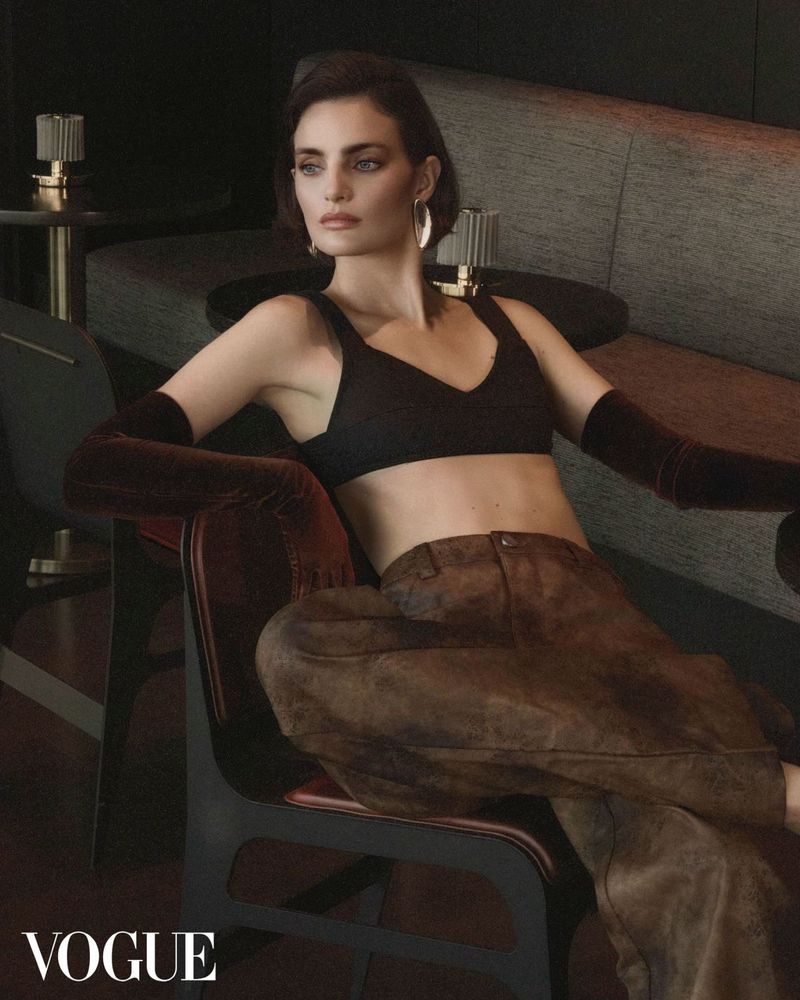 Elegant woman in a dark crop top and brown pants sitting in a modern chair, posing in an indoor setting, with the Vogue logo.