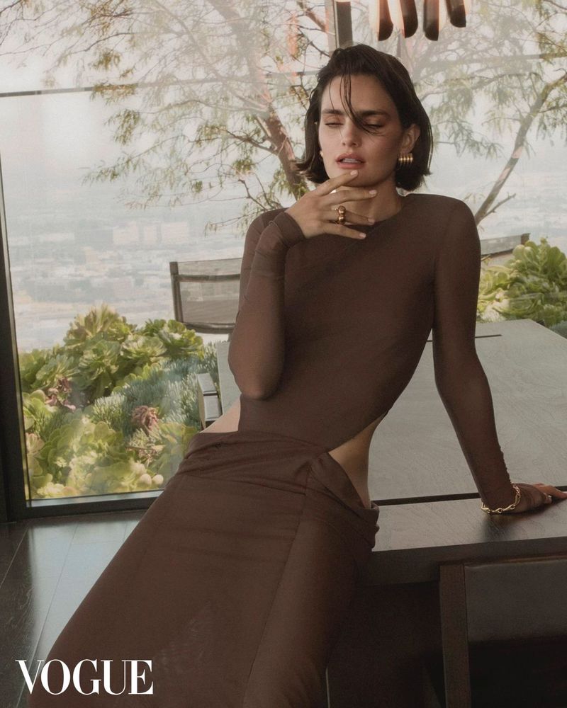 A woman with short dark hair, wearing a long-sleeved brown dress with a side cut-out, leans against a table in a modern home interior.