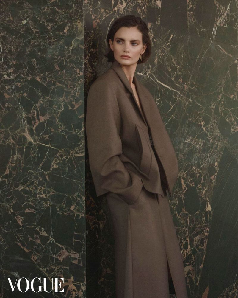 A fashion editorial shot showcasing a brunette woman in a gray suit against a green marble wall, featuring the 'Vogue' magazine logo.