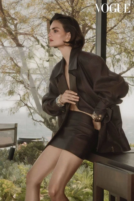 Brunette woman in a black leather jacket and miniskirt poses near a window with outdoor greenery and a glimpse of the sea.