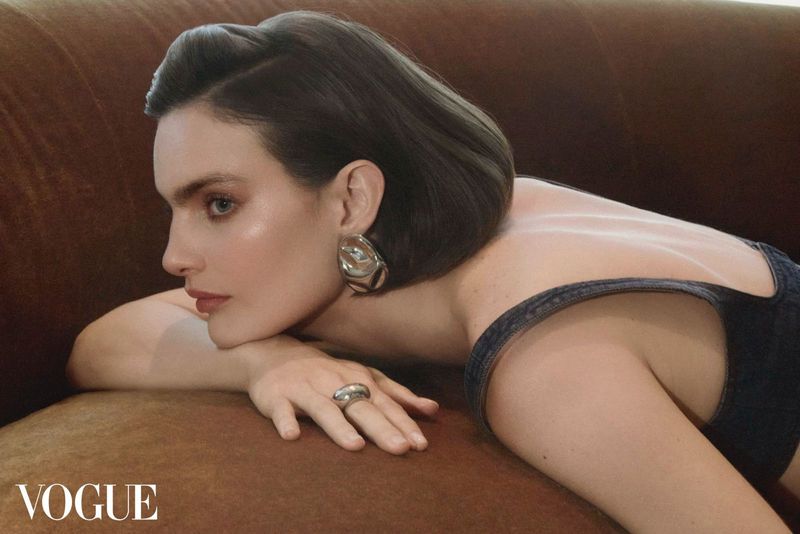 Woman poses on brown sofa wearing statement jewelry in close-up studio shot for fashion editorial with Vogue logo at lower left.
