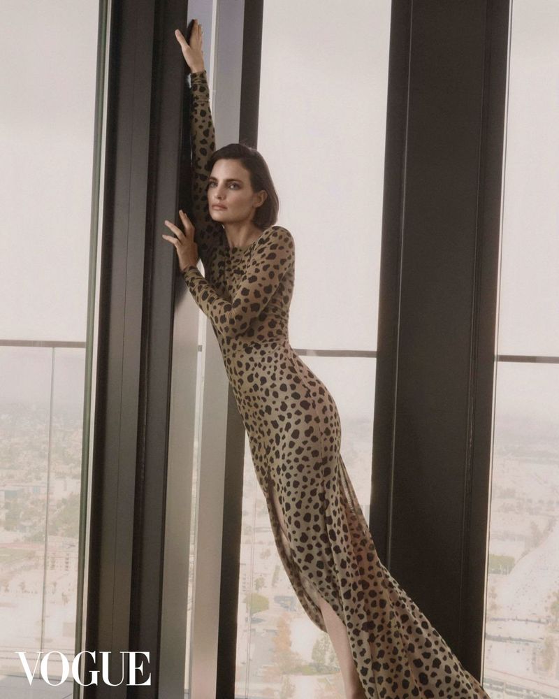 Fashion shot of a single adult woman in a long cheetah-print dress leaning against a dark window frame, with a city view in background.