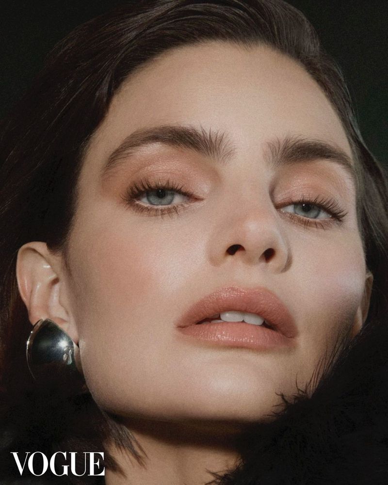 Close-up portrait of a model with fair skin, blue eyes, and dark hair, adorned with a silver earring, for a Vogue magazine beauty shot.