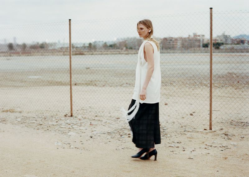 Fashion shot of a blonde adult woman in a white sleeveless top and dark culottes walking in a gravel field near a chain link fence.