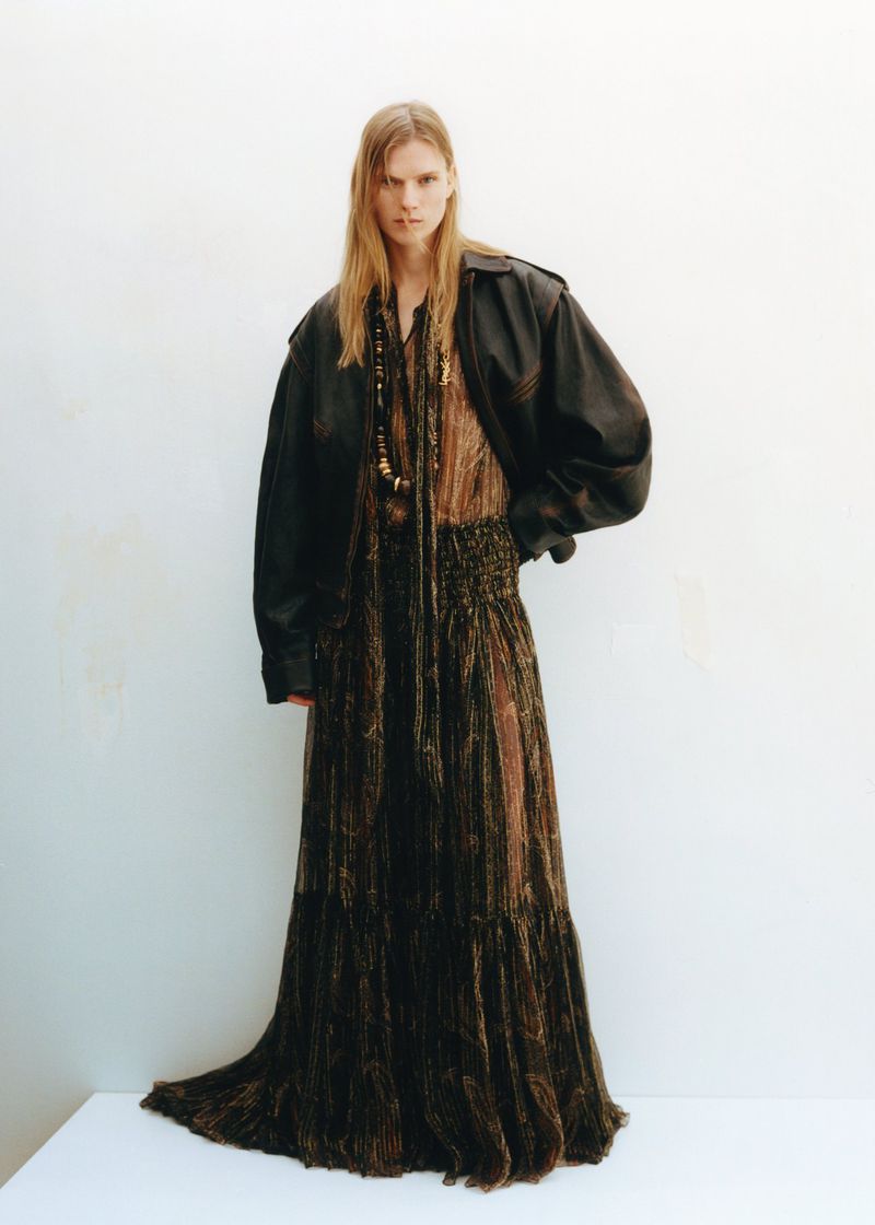 A full-length shot of an adult woman in a brown dress and black jacket, standing on a white surface against a white backdrop.