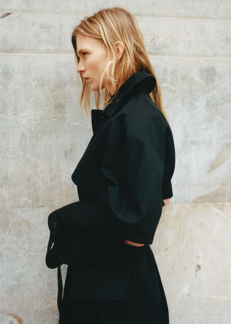 Fashion shot of a blonde woman in a black coat posing against a weathered wall, emphasizing minimalist style and neutral tones.