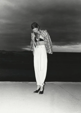 Monochrome fashion shot of a woman in snake-print jacket, bra top, and high-waisted pants against a dark horizon under stormy clouds.