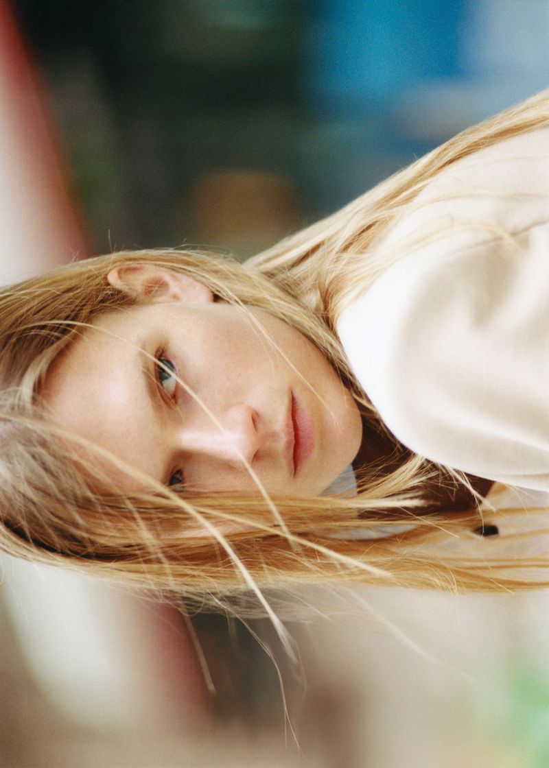 Close-up portrait of a blonde model with hair across her face in a creamy top for Puss Puss Magazine