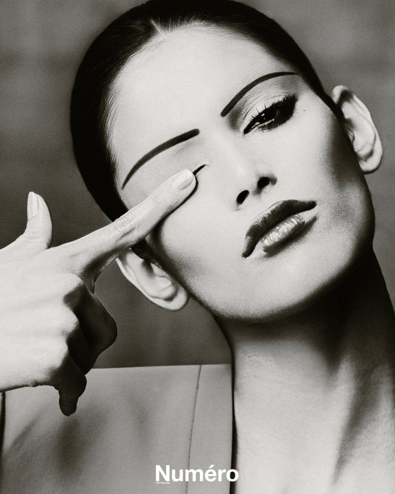 Black and white close-up of a woman with striking makeup, covering one eye with her finger, evoking a dramatic fashion editorial.