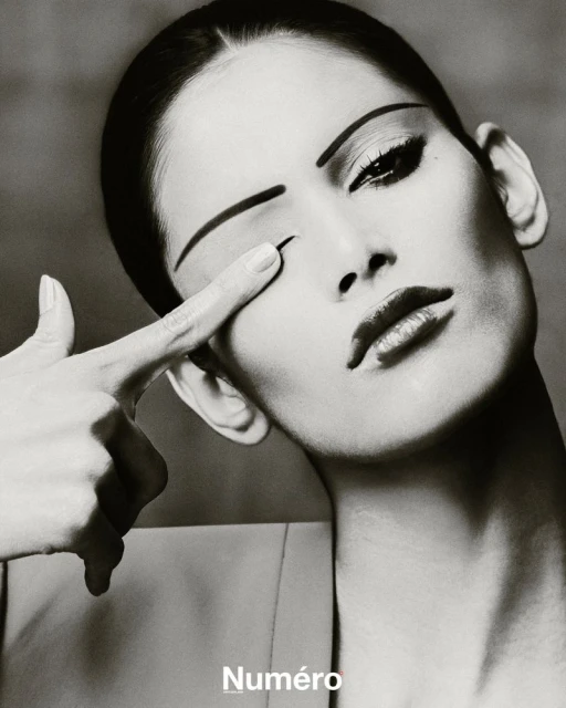 Black and white close-up of a woman with striking makeup, covering one eye with her finger, evoking a dramatic fashion editorial.