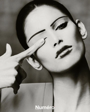 Black and white close-up of a woman with striking makeup, covering one eye with her finger, evoking a dramatic fashion editorial.
