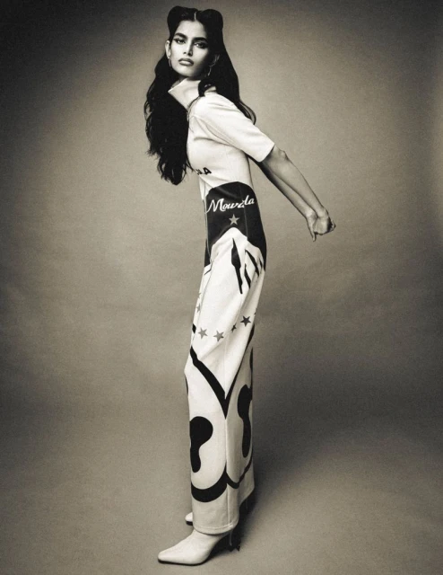 Studio portrait of a tall, slender woman in printed white top and pants, with heeled boots, in a fashion editorial pose.