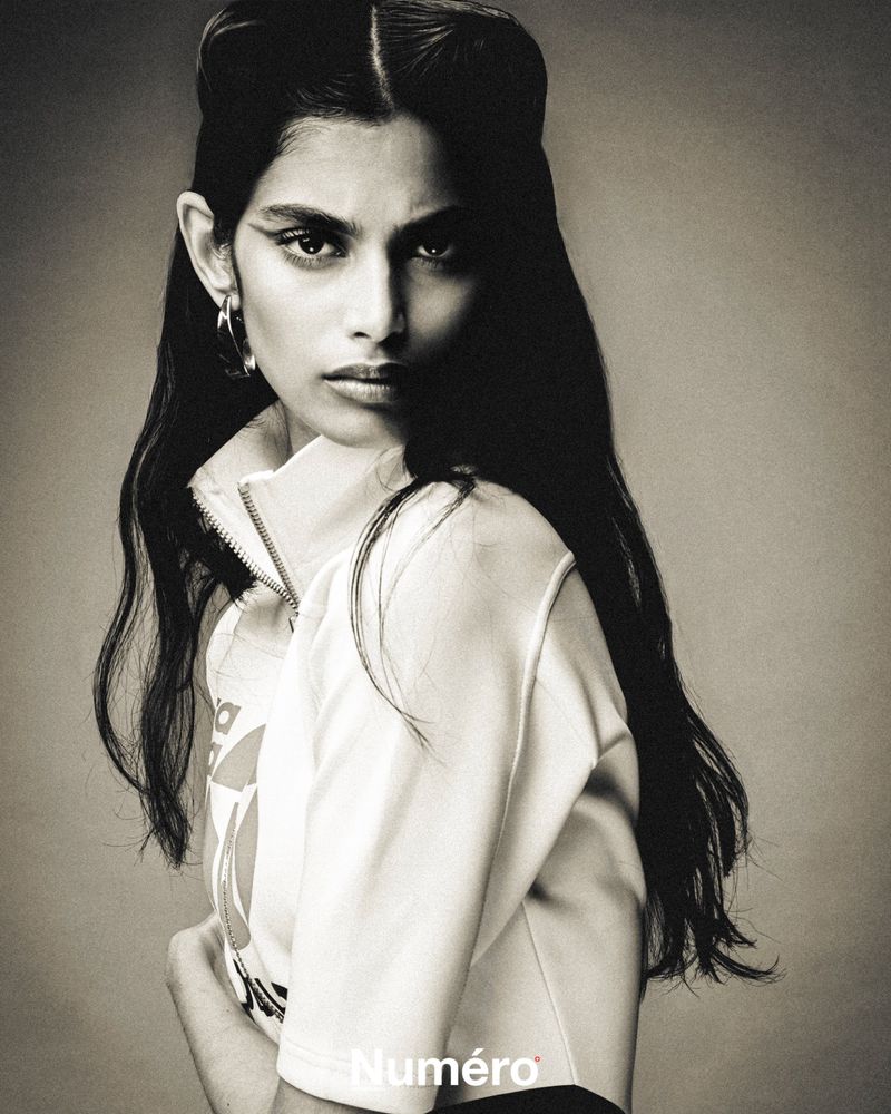 Monochrome portrait of an adult female with dark hair wearing a jacket, hoop earrings, and strong eyeliner in a photo studio.