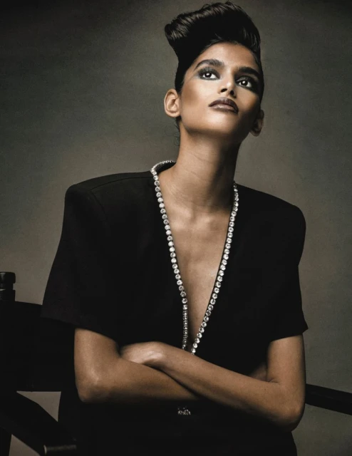 Close-up fashion portrait of a slim adult woman with crossed arms, wearing a black top and a diamond necklace, looking up against gray background.