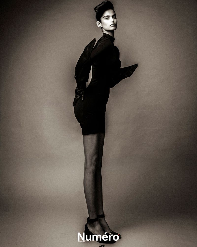 A striking sepia-toned fashion portrait of a slender woman in a black dress, styled with puffed sleeves and ankle-strap heels, in studio.