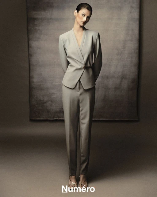 Elegant woman in a light grey suit with a plunging neckline poses in front of a grey backdrop, showcasing a modern and stylish look.