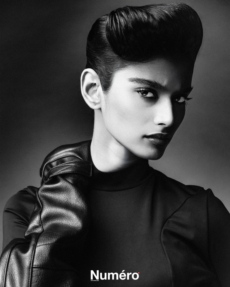 Black and white portrait of a woman with an elegant pompadour hairstyle, wearing black leather gloves and a black top against a dark backdrop.