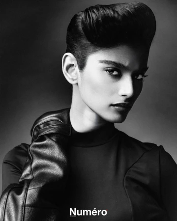Black and white portrait of a woman with an elegant pompadour hairstyle, wearing black leather gloves and a black top against a dark backdrop.