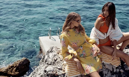 Two models posing on rocky shore beside blue water for Zimmermann Resort Swim, wearing floral dress and orange swimwear.