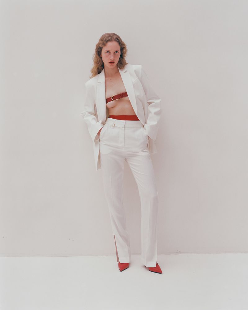 Fashion shot of a female model with curly hair wearing a white suit with a red bandeau and matching heels against a white background.