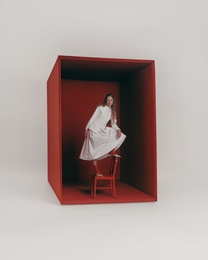 A young woman in a white suit strikes a dynamic pose on a red chair inside a vivid red box against a clean white background.