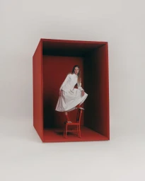 Model in white dress standing on red chair inside a large red box for Elle Greece Spring cherries editorial