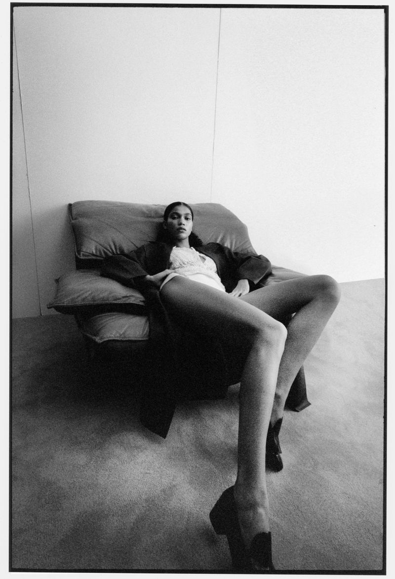 Elegant woman in repose on a chair wearing a robe and heels. The black-and-white portrait is set against a stark white wall.